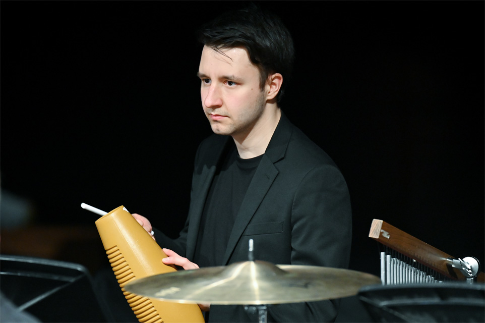 Male student, wearing smart attire, looking at the conductor, playing Latin instruments in a jazz performance.
